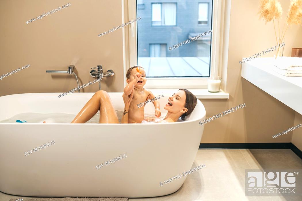 Stock Photo: Merry young female parent supporting the child with a toy seated on her in the bath.