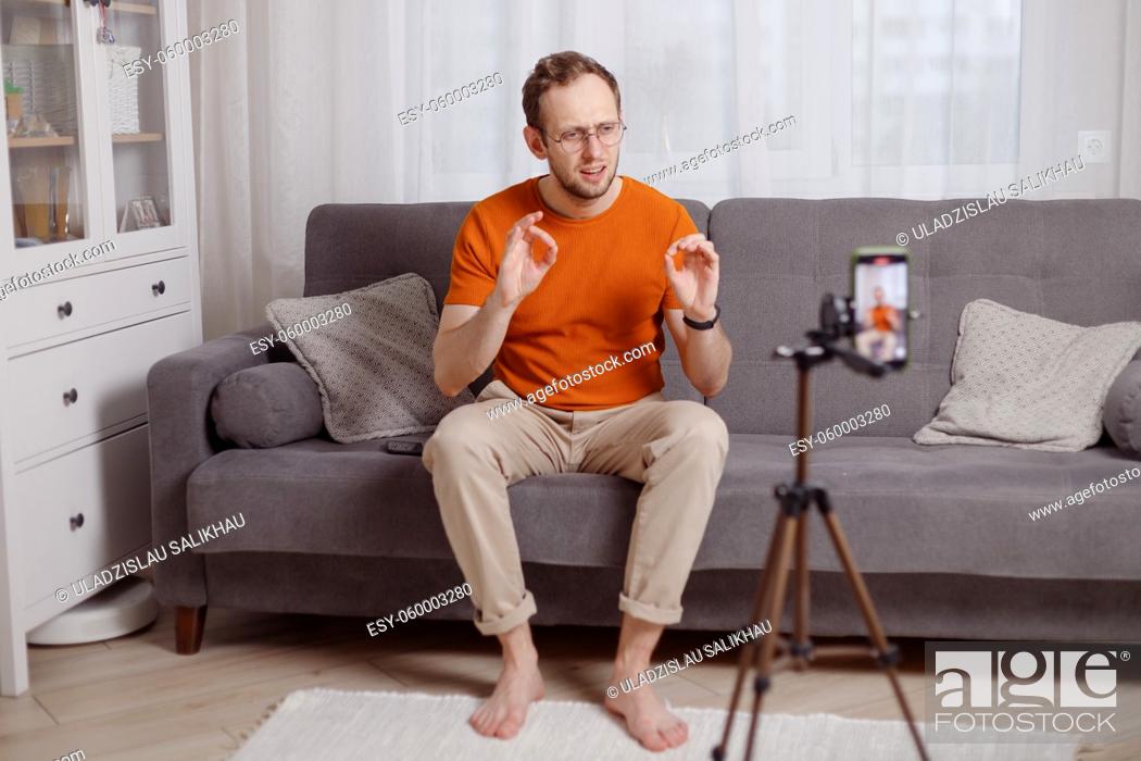 Stock Photo: Hipster man blogger recording vlog on mobile phone sit on couch in living room and explain and gesturing with hands.