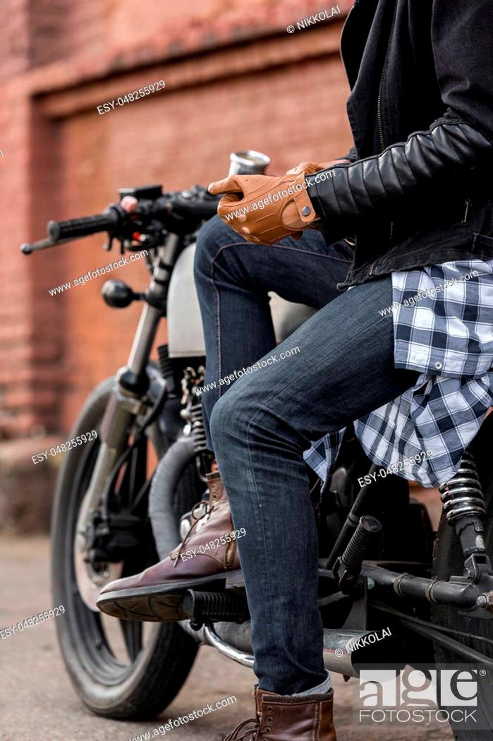 Stock Photo: Close up of a handsome rider biker man hand puts on a leather glove while sit on classic style cafe racer motorcycle. Bike custom made in vintage garage.