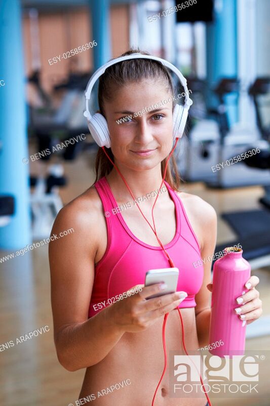 Stock Photo: young healthy woman drinking water in fitness gym while sitting on pilates ball and listening music on headphones from smartphone.