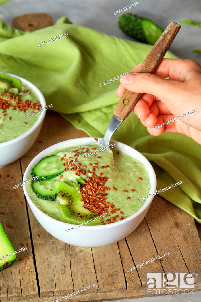 Photo de stock: Freshly prepared smoothies. A woman's hand with a spoon takes a green smoothie of cucumber, avocado, kiwi and flax seeds against a wooden board.