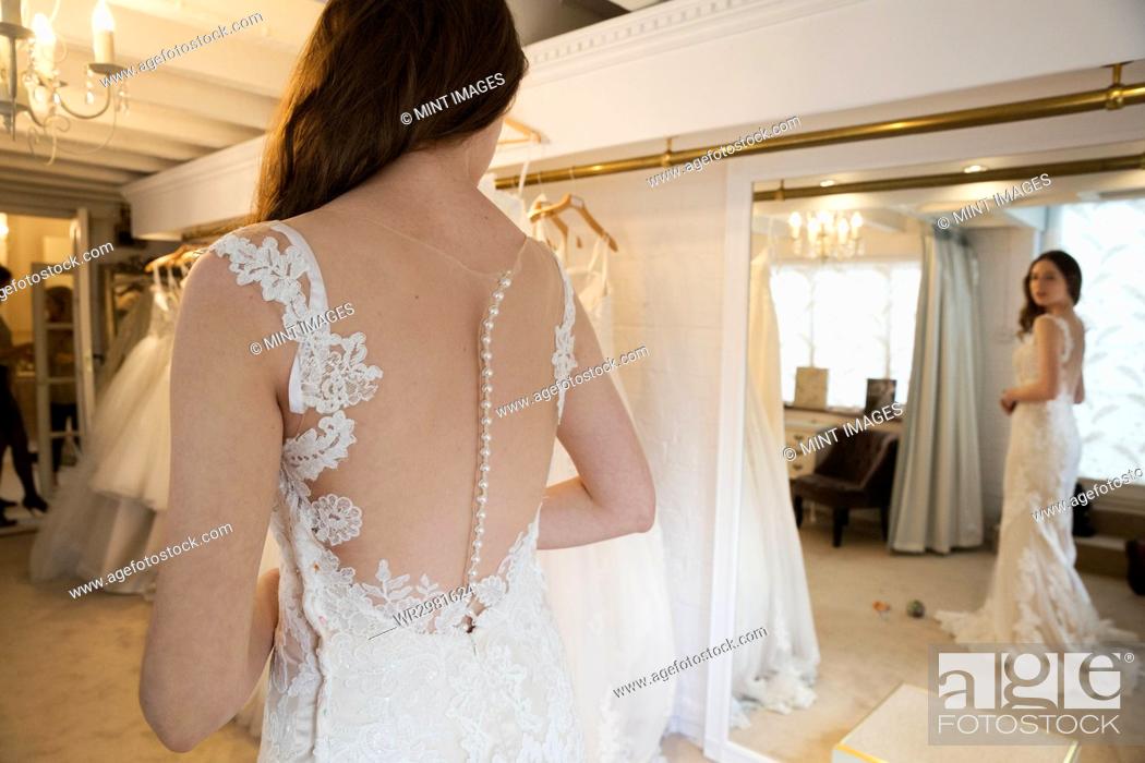 Imagen: A young woman in a full length white wedding dress, looking at her reflection in the mirror in a bridal boutique.