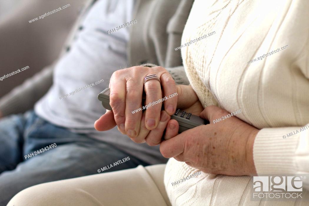 Stock Photo: Elderly woman holding television remote with both of her hands. Aged grandma wearing white sweater holding tv remote with her husband next to her.