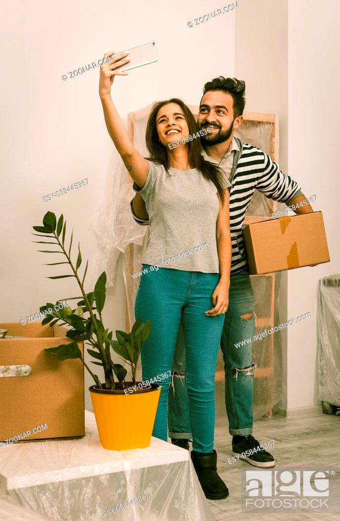 Stock Photo: Man and woman in casual clothes standing in new home after relocation, forwarding to new life. Couple making selfie shot in living room witn new furniture.