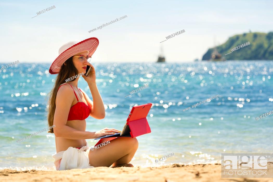 Stock Photo: Fashionable young woman talking on mobile phone while using a tablet on the beach in a sunny day of summer in Flores Island, Indonesia.