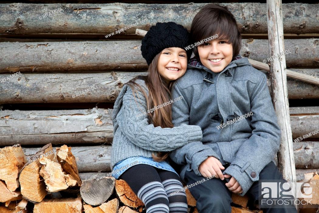 Stock Photo: Germany, Huglfing, Girl and boy sitting on stack of firewood, smiling, portrait.