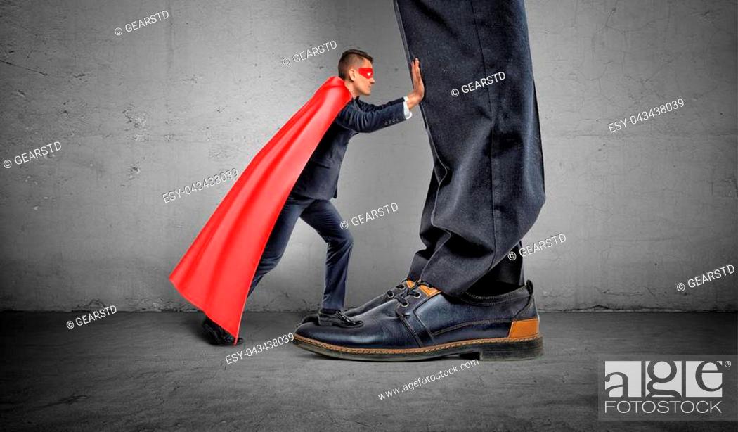 Stock Photo: A small businessman in a superhero cape trying to push away giant man legs. Uneven competition. Best performance. Business problems.