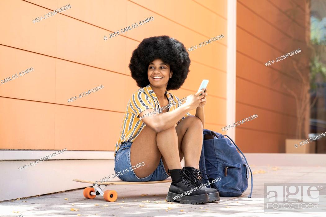 Stock Photo: Woman with backpack holding mobile phone while sitting on skateboard.