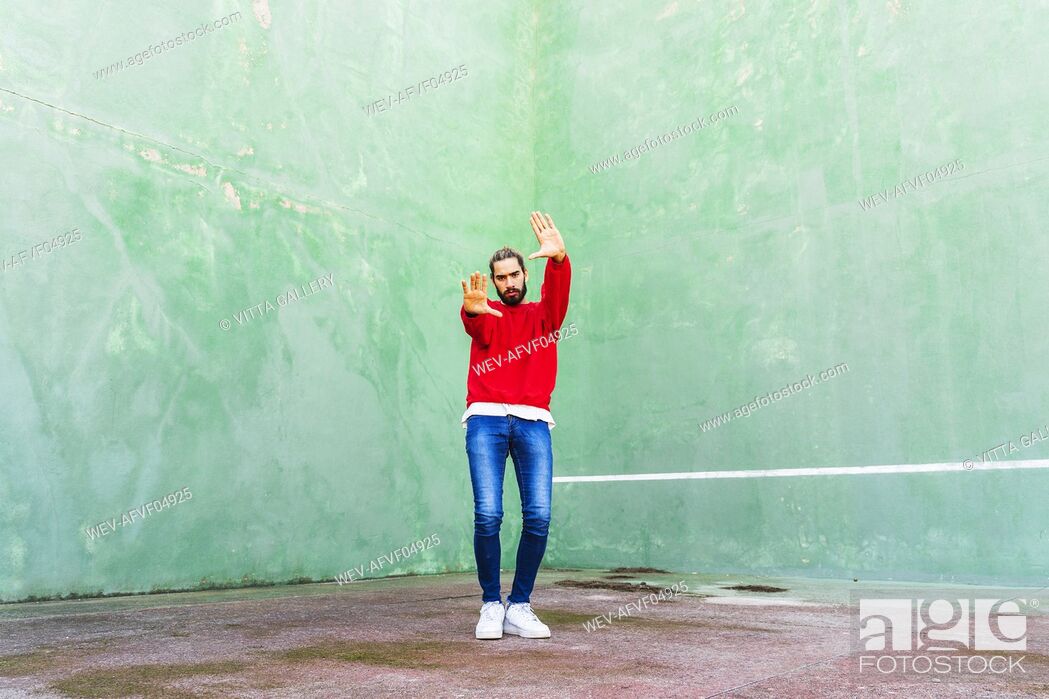Stock Photo: Portrait of serious young man wearing red sweatshirt raising hands in front of green wall.