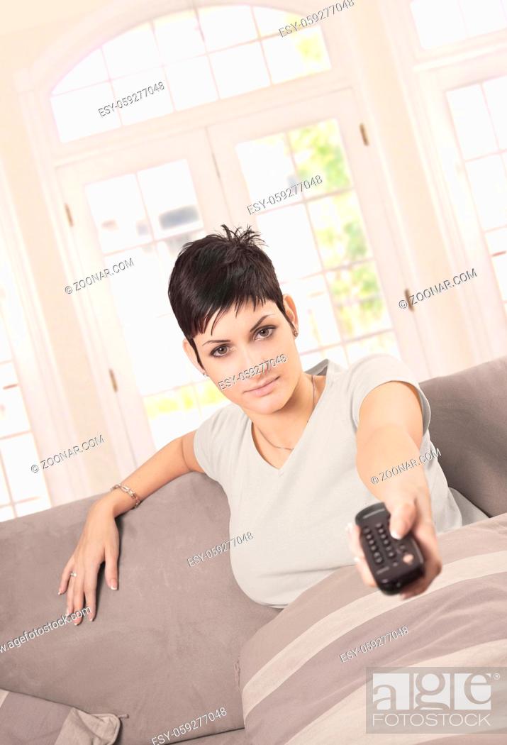 Stock Photo: Young woman is watching TV at home in the living room with a remote controller in hand.