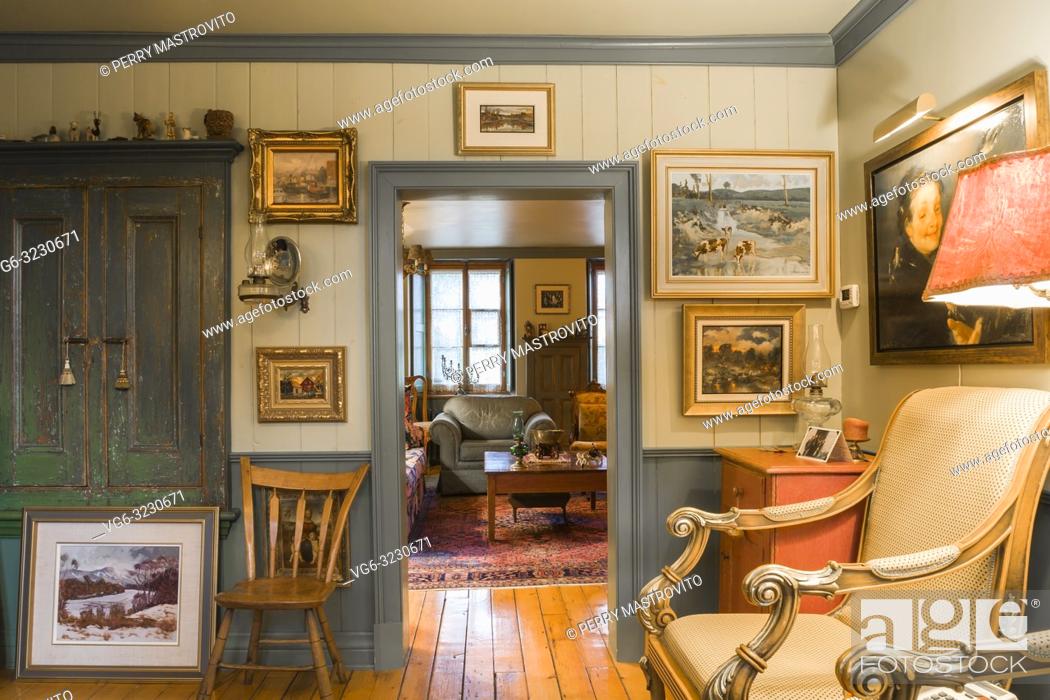 Stock Photo: Old wooden chair and upholstered armchair in parlour room decorated with several paintings and doorway leading to living room inside an old circa 1805 Canadiana.