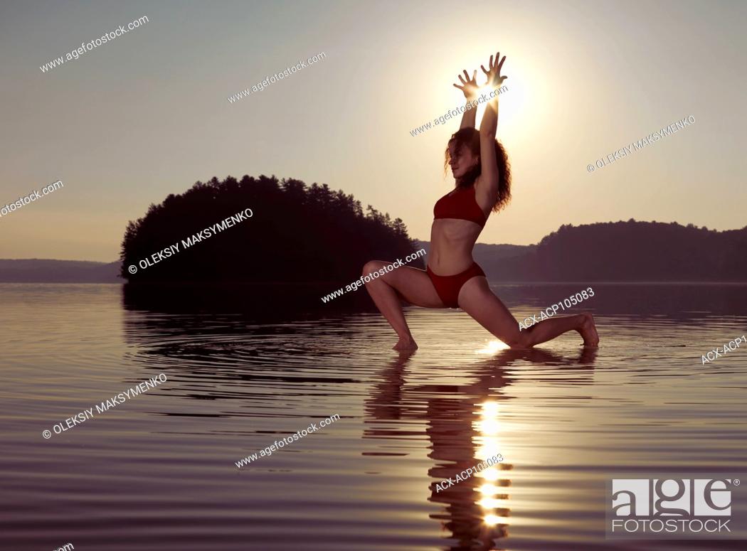 Stock Photo: Young woman practicing Hatha yoga artistic variation of Low Lunge pose on a floating platform in water on the lake during misty sunrise in the morning.
