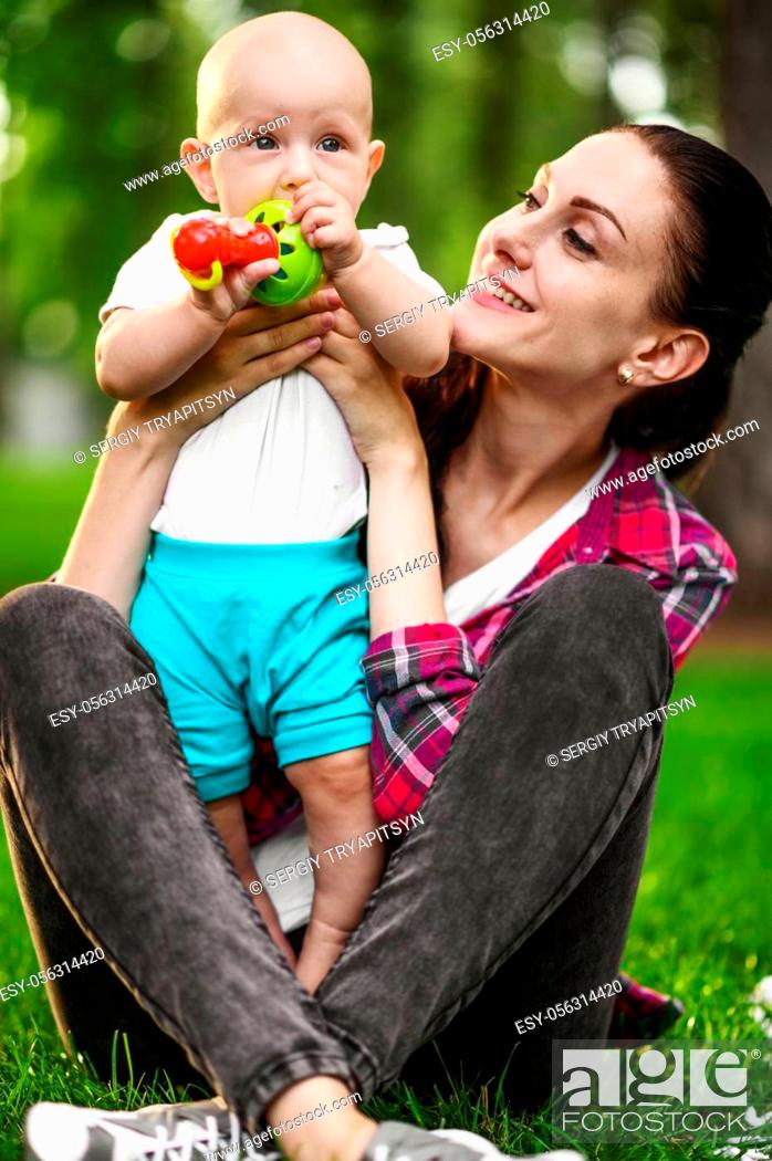 Imagen: Portrait of mother and her little baby are sitting on the grass in summer park. Mom with male kid leisures on lawn, picnic with child on plaid in the forest.
