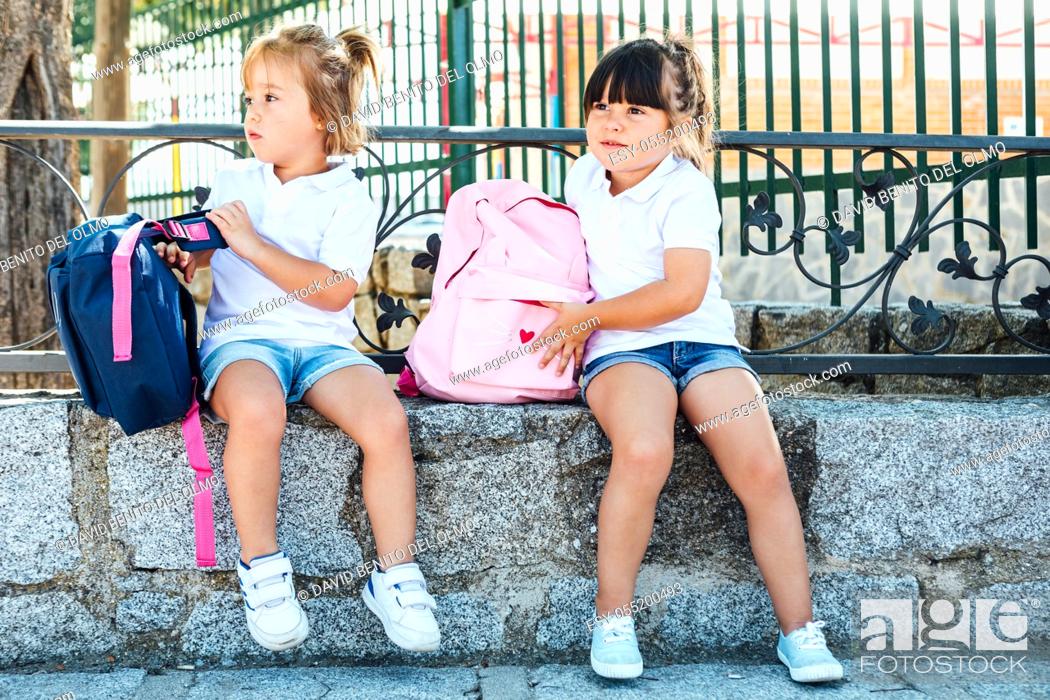 Stock Photo: A black-haired girl wearing a pink backpack and a blonde-haired girl wearing a blue backpack next to the school. School concept.