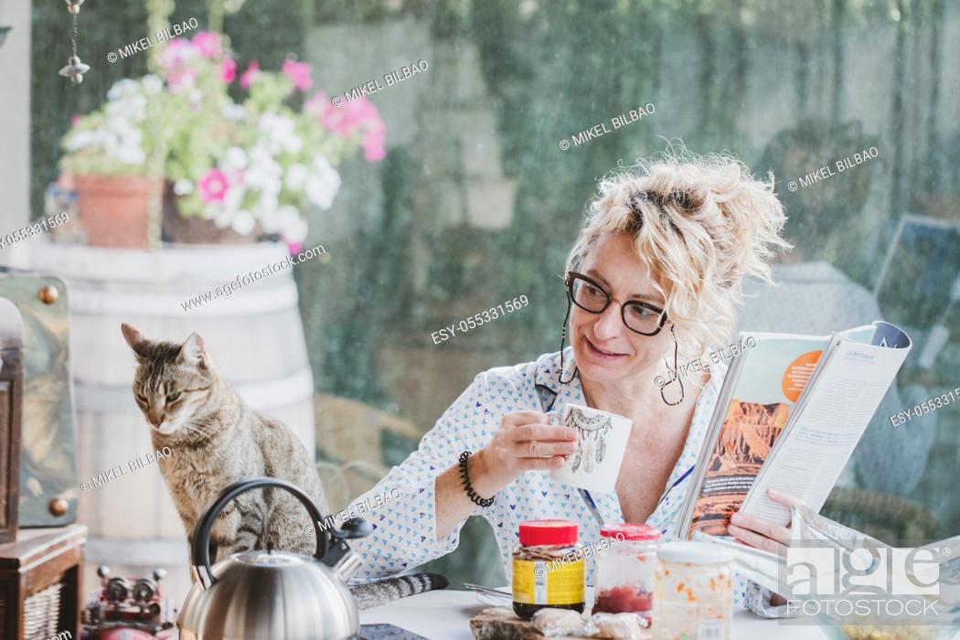 Stock Photo: Blonde young mature woman with glasses in pyjamas at home in breakfast time, reading a magazine and having a cup of coffee and cat.