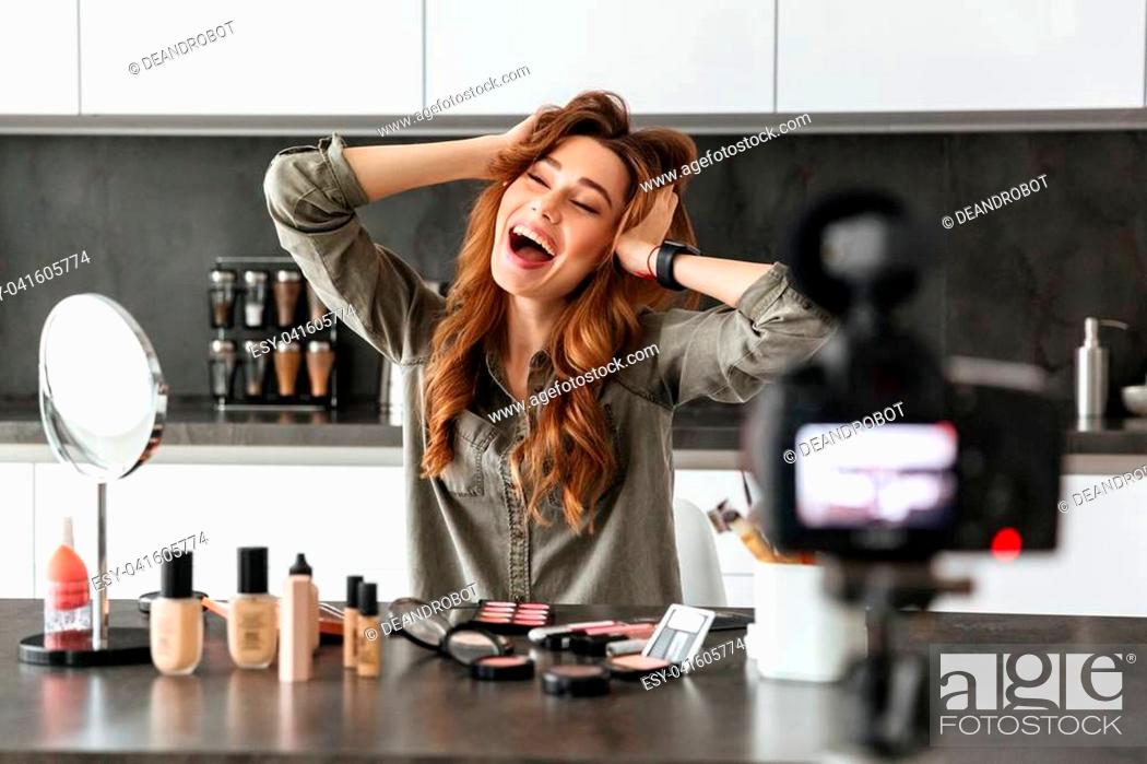 Photo de stock: Cheerful young girl recording her video blog episode about new cosmetic products while sitting at the kitchen table at home and applying make-up and showing ok.