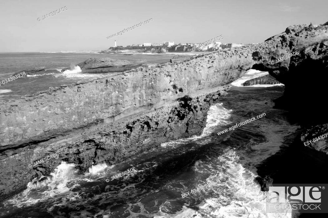 Stock Photo: Black and white photo of sandstone rock arch in the sea off Biarritz, with lighthouse in distance.