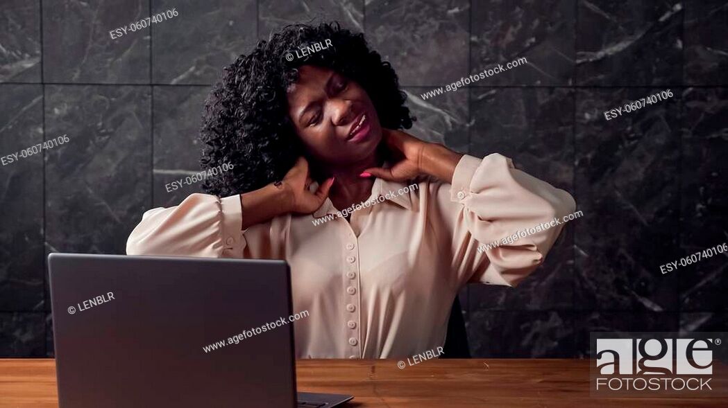 Stock Photo: Bored Afro-american secretary with curly hair stretches back sitting at brown wooden table with grey laptop by marble wall in empty office.