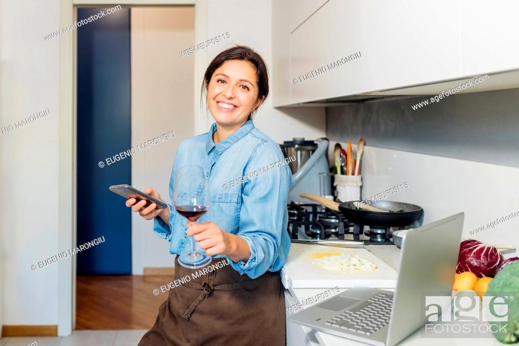 Stock Photo: Woman holding wineglass and smart phone leaning on kitchen counter at home.