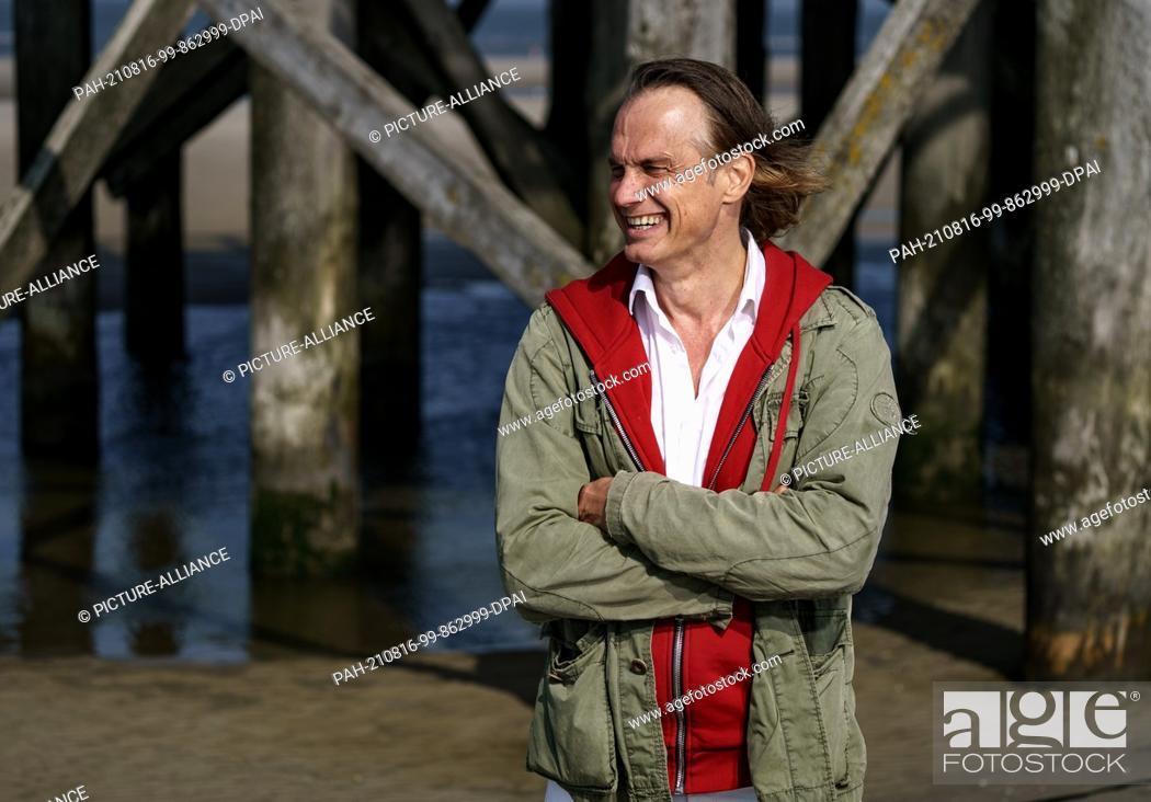 Stock Photo: 12 August 2021, Schleswig-Holstein, Sankt Peter-Ording: Actor Ralf Bauer stands on the beach of Sankt Peter-Ording in front of one of the traditional pile.