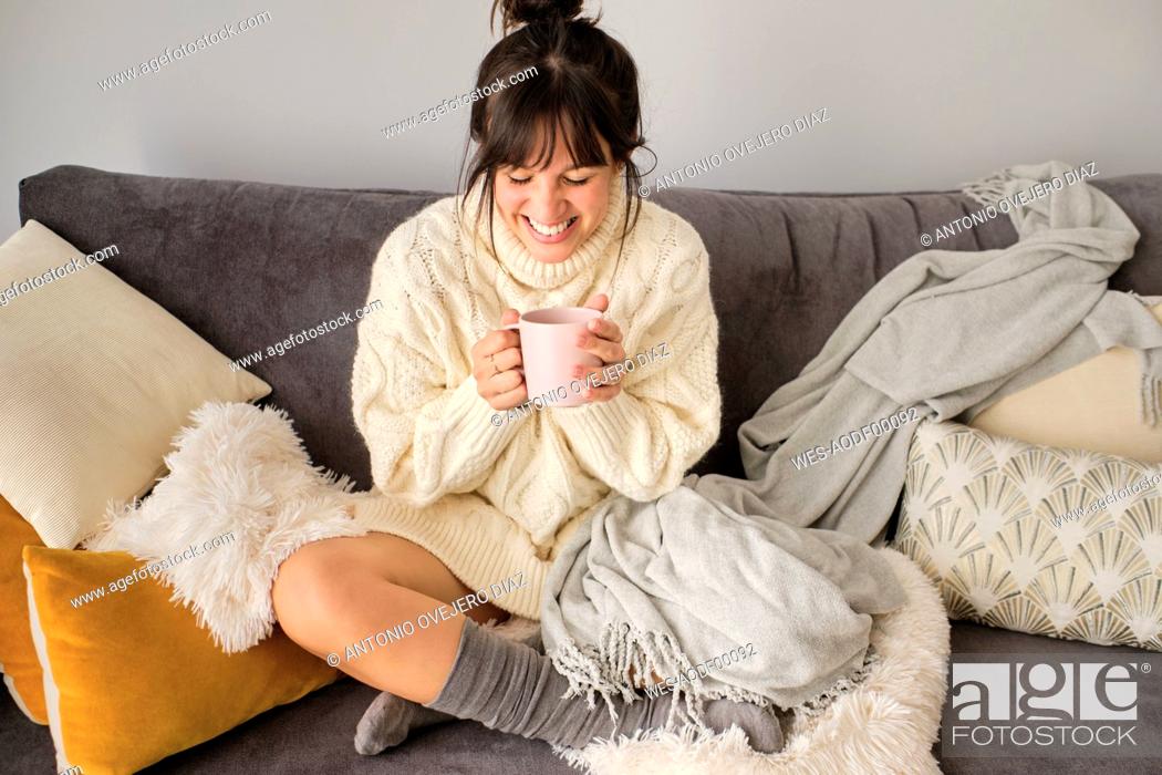 Stock Photo: Happy woman having coffee in mug while sitting on sofa at home during winter.