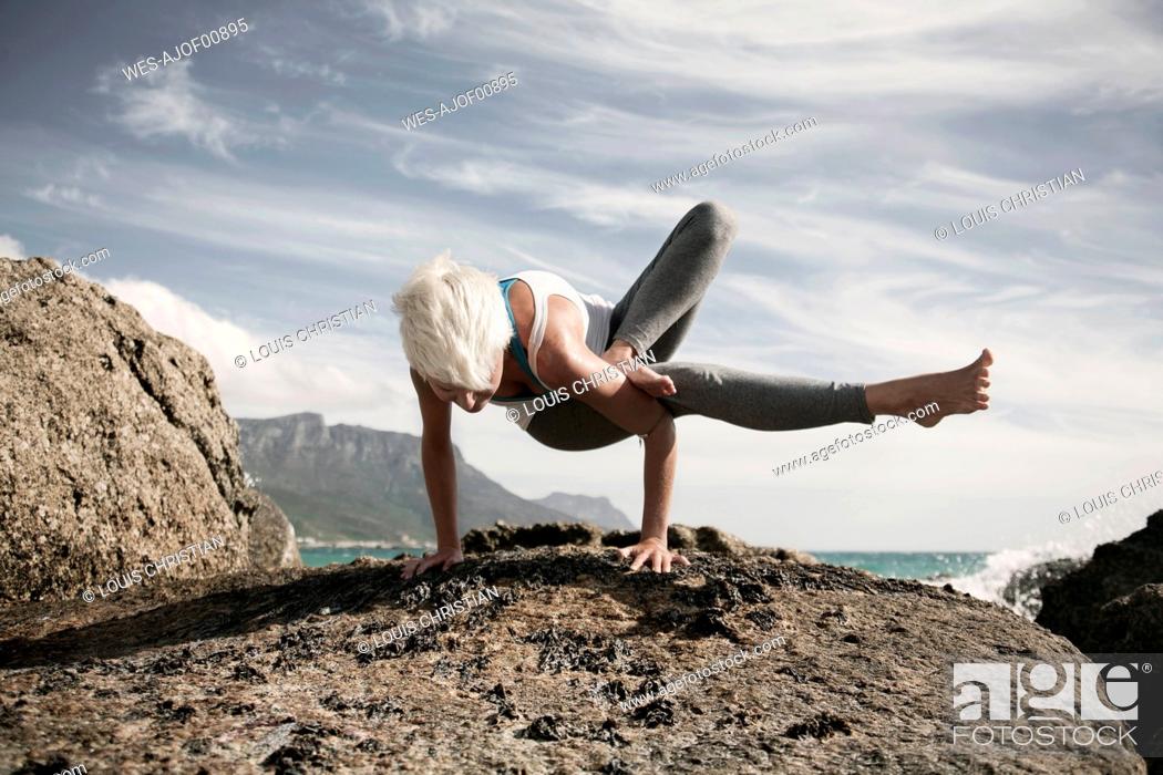 Stock Photo: Blond woman practicing Astavakrasana on rock formation against sky.