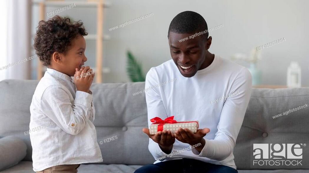 Imagen: Shocked happy african american man looking at wrapped box gift from funny mixed race cute preschool son, sitting at sofa in living room at home.