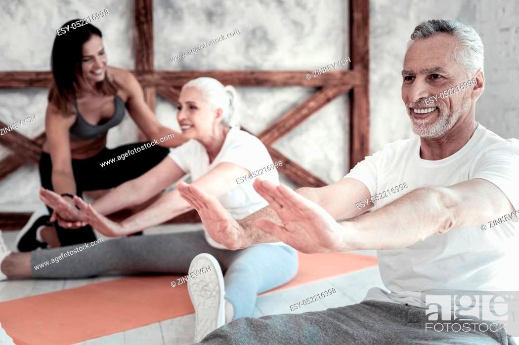 Stock Photo: Feeling so much better. Selective focus on a joyful senior man beaming while sitting on a mat and stretching his body during a group training session.