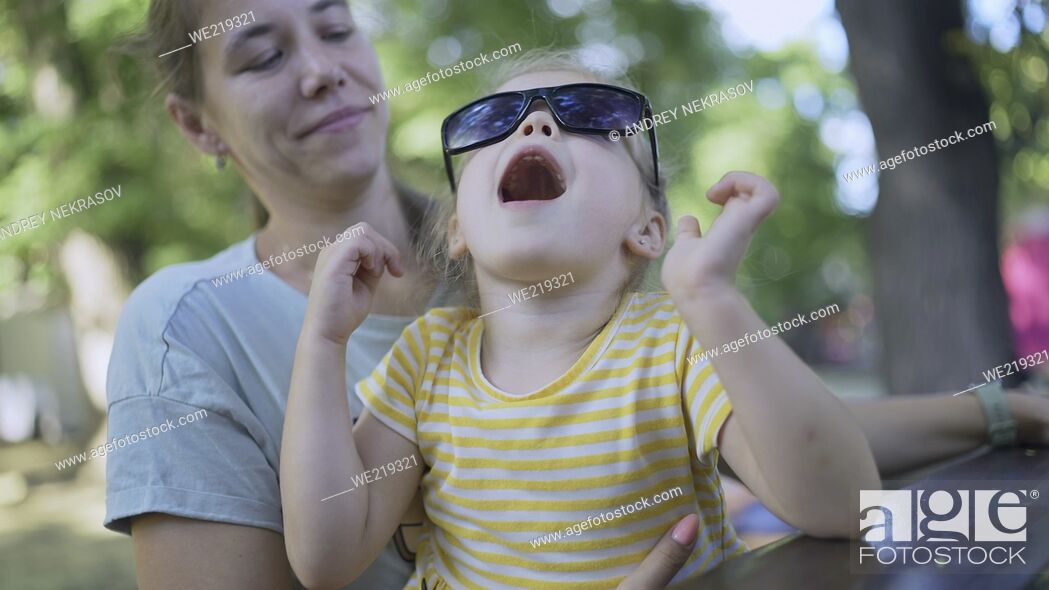 Stock Photo: Closeup of mom puts her sunglasses on cute little girl sitting in the park. Close-up of mother and daughter playing while sitting on a park bench in a street.