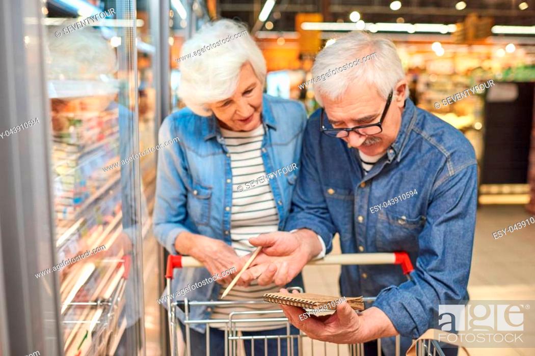 Stock Photo: Portrait of smiling senior couple grocery shopping in supermarket, reading shopping list leaning on cart standing by freezers.