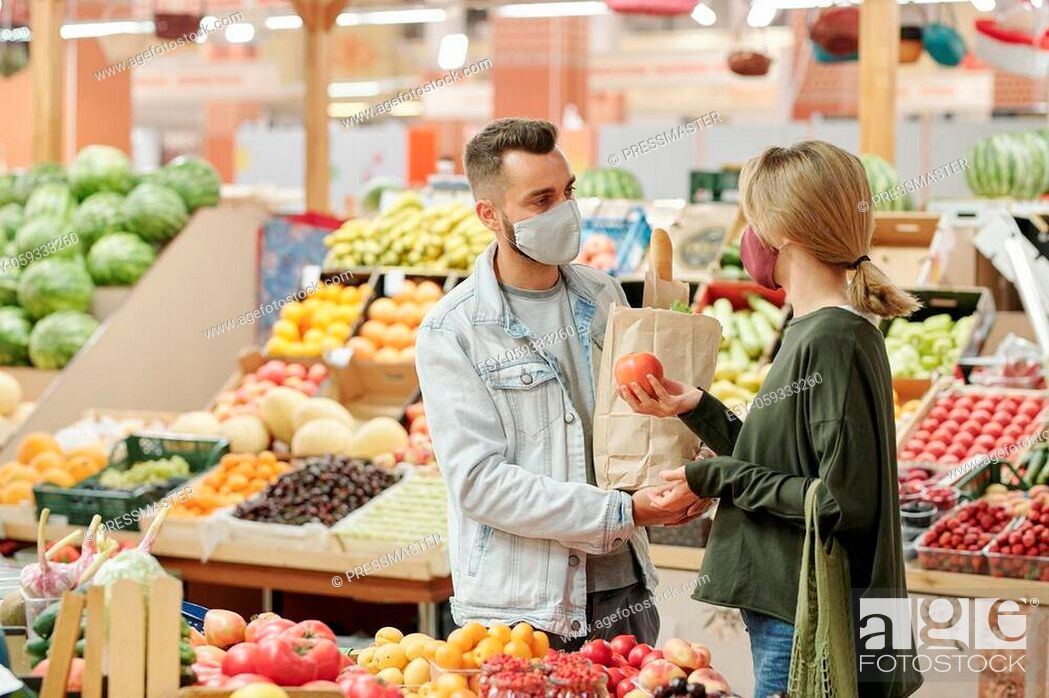Stock Photo: Young couple in cloth masks standing at counter and buying organic food at market: girl choosing tomatoes.