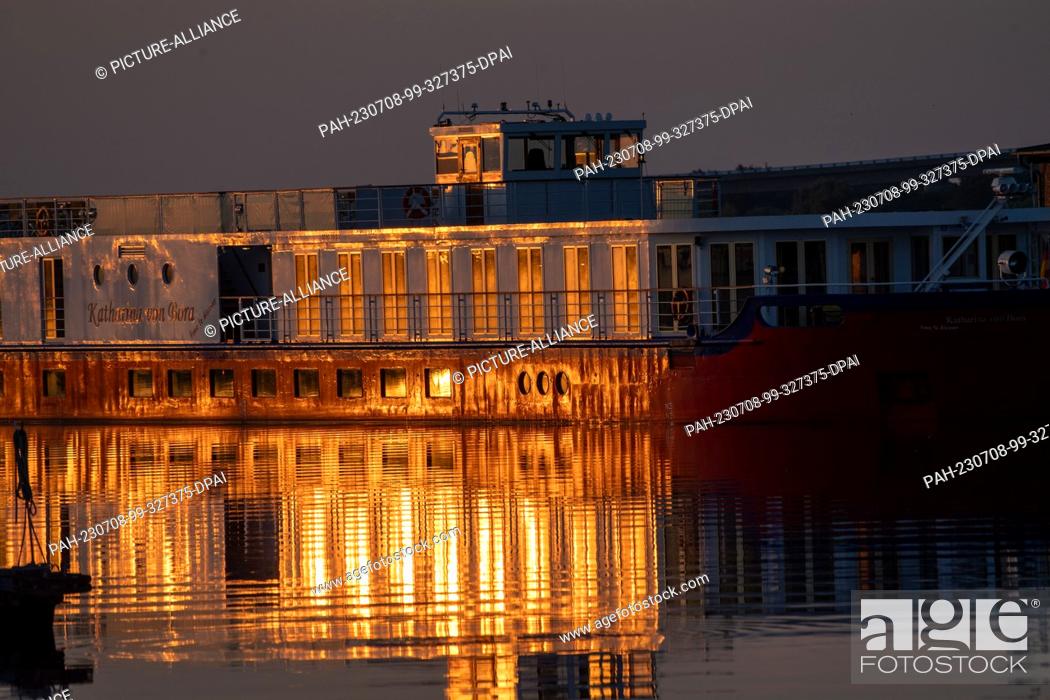 Stock Photo: 08 July 2023, Mecklenburg-Western Pomerania, Stralsund: The rising sun bathes a river cruise ship in a warm light in the harbor.