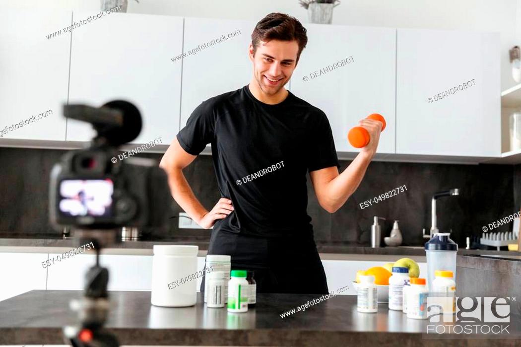 Stock Photo: Happy young man filming his video blog episode about healthy food additives while standing at the kitchen table and exercising with a dumbbell.